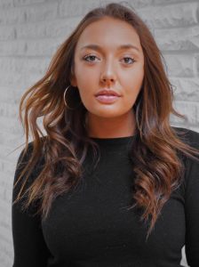 A woman with long, wavy brown hair wearing a black top and large hoop earrings stands in front of a white brick wall, looking directly at the camera with a neutral expression.