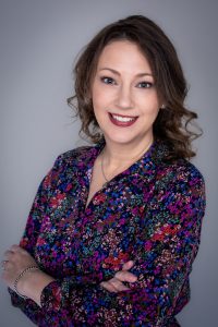 Smiling woman with wavy brown hair, wearing a colorful floral blouse and silver jewelry, standing with arms crossed against a plain gray background.