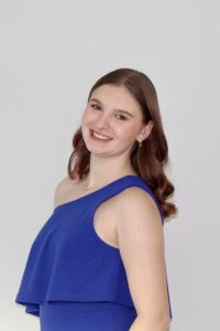 A young woman with wavy brown hair smiles at the camera. She is wearing a sleeveless, off-the-shoulder royal blue dress and small hoop earrings. The background is plain and light grey.