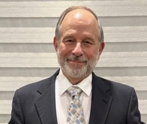 A middle-aged man with a beard and receding hairline is wearing a suit, white shirt, and patterned tie. He stands in front of a light-colored, horizontally-striped background, smiling slightly.