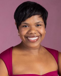 A woman with short black hair, wearing gold hoop earrings and a sleeveless magenta top, smiles at the camera against a pink background.