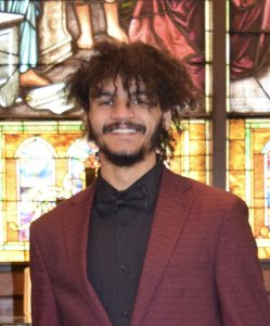 A young man with curly hair and a beard, wearing a maroon suit jacket and black bow tie, stands smiling in front of colorful stained glass windows. A young man with curly hair and a beard, wearing a maroon suit jacket and black bow tie, stands smiling in front of colorful stained glass windows.