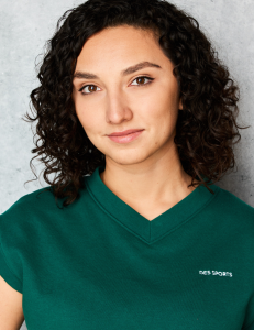 A woman with curly dark hair and brown eyes wears a green top with DES SPORTS written on it, standing against a light gray textured background, looking confidently at the camera. A woman with curly dark hair and brown eyes wears a green top with DES SPORTS written on it, standing against a light gray textured background, looking confidently at the camera.