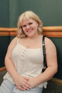 A smiling person with blonde hair sits against a dark green wall, wearing a light sleeveless top, jeans, and a black shoulder bag. Their hands are folded in their lap, and they have light purple nails and a ring.