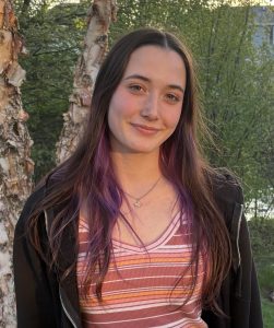 A young woman with long brown hair and purple streaks stands outdoors, smiling softly. She wears a pink striped top and a black jacket. Sunlight filters through trees in the background. A young woman with long brown hair and purple streaks stands outdoors, smiling softly. She wears a pink striped top and a black jacket. Sunlight filters through trees in the background.