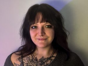 A person with long dark hair, bangs, and facial piercings smiles slightly at the camera. They have visible tattoos on their chest and are wearing a dark sweater and layered necklaces, standing against a plain light background.