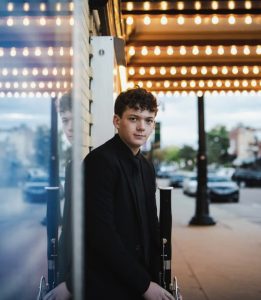 A young man in a black suit stands by a window, his reflection visible. Behind him, a sidewalk and city street are lit by rows of bright marquee lights overhead. The scene is calm and slightly overcast.