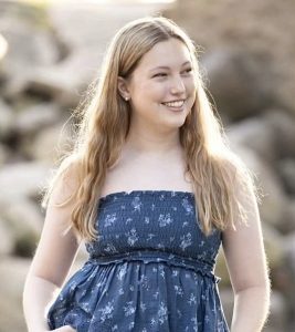 A young woman with long blonde hair wearing a blue, sleeveless dress with a floral pattern stands outdoors, smiling. The background is blurred with natural stones and sunlight.