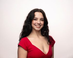 A young woman with long wavy dark hair smiles at the camera. She is wearing a bright red dress with a deep neckline, standing against a plain light background.