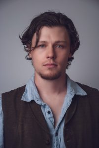 A young man with wavy brown hair and blue eyes looks directly at the camera. He is wearing a light blue button-up shirt and a dark vest, standing against a plain gray background.