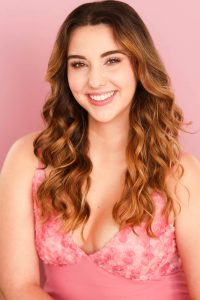 A young woman with long, wavy brown hair smiles warmly. She wears a sleeveless pink dress with floral details and poses in front of a light pink background. A young woman with long, wavy brown hair smiles warmly. She wears a sleeveless pink dress with floral details and poses in front of a light pink background.
