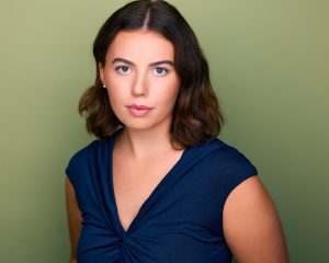 A young woman with shoulder-length brown hair and light skin looks directly at the camera, wearing a navy blue top, standing against a plain light green background. A young woman with shoulder-length brown hair and light skin looks directly at the camera, wearing a navy blue top, standing against a plain light green background.