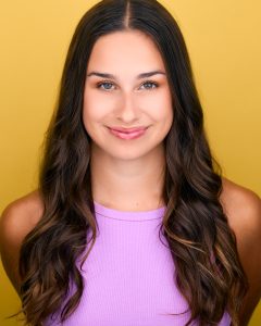 A young woman with long, wavy brown hair smiles at the camera. She is wearing a sleeveless lavender top and stands in front of a bright yellow background. A young woman with long, wavy brown hair smiles at the camera. She is wearing a sleeveless lavender top and stands in front of a bright yellow background.