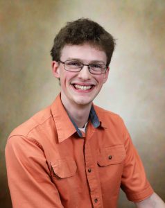 A young man with short curly hair and glasses, wearing an orange button-up shirt, smiles at the camera against a neutral gradient background. A young man with short curly hair and glasses, wearing an orange button-up shirt, smiles at the camera against a neutral gradient background.
