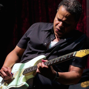 A man wearing a black shirt passionately plays an electric guitar on stage, with a dark red curtain in the background.