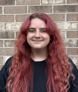 A person with long, wavy pink hair smiles while standing in front of a brick wall, wearing a black shirt. A person with long, wavy pink hair smiles while standing in front of a brick wall, wearing a black shirt.