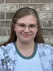 A person with long, straight light brown hair, glasses, and a headband stands in front of a brick wall, wearing a patterned shirt over a white tee with a green collar, smiling slightly. A person with long, straight light brown hair, glasses, and a headband stands in front of a brick wall, wearing a patterned shirt over a white tee with a green collar, smiling slightly.