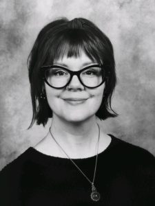 Black-and-white portrait of a person with short hair, wearing large dark glasses, a dark top, and a round pendant necklace, smiling at the camera with a neutral background.