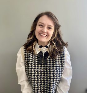 A woman with wavy brown hair smiles, wearing a houndstooth sweater vest over a white shirt with a decorative brooch and black bow, standing against a plain gray background. A woman with wavy brown hair smiles, wearing a houndstooth sweater vest over a white shirt with a decorative brooch and black bow, standing against a plain gray background.
