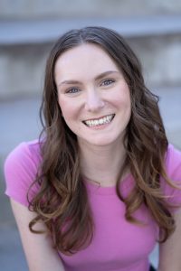 A woman with long brown hair and blue eyes smiles at the camera. She is wearing a pink top and sitting outdoors in front of concrete steps. A woman with long brown hair and blue eyes smiles at the camera. She is wearing a pink top and sitting outdoors in front of concrete steps.