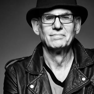 A man wearing glasses, a black hat, and a leather jacket looks at the camera. The black and white photo has a neutral background and dramatic lighting, highlighting his facial features and clothing.