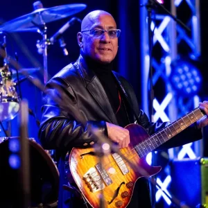 A bald man wearing glasses and a black leather jacket plays an electric bass guitar onstage in front of drums and blue stage lights.