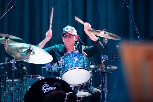 A drummer wearing a cap and glasses plays a blue drum kit on stage. A drummer wearing a cap and glasses plays a blue drum kit on stage.