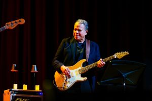 A man in a dark suit playing an electric guitar on stage. A man in a dark suit playing an electric guitar on stage.