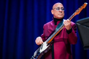 A bald man with glasses wearing a maroon jacket plays a bass guitar on stage in front of a blue curtain. A bald man with glasses wearing a maroon jacket plays a bass guitar on stage in front of a blue curtain.